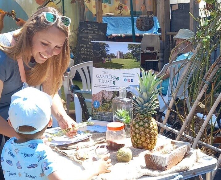 An adult woman and child leant over a table in a sunny garden. The table displays a selection of everyday objects such as a pineapple, seeds, brick and tennis ball.