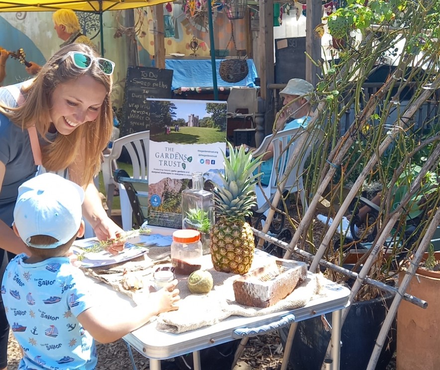 An adult woman and child leant over a table in a sunny garden. The table displays a selection of everyday objects such as a pineapple, seeds, brick and tennis ball.