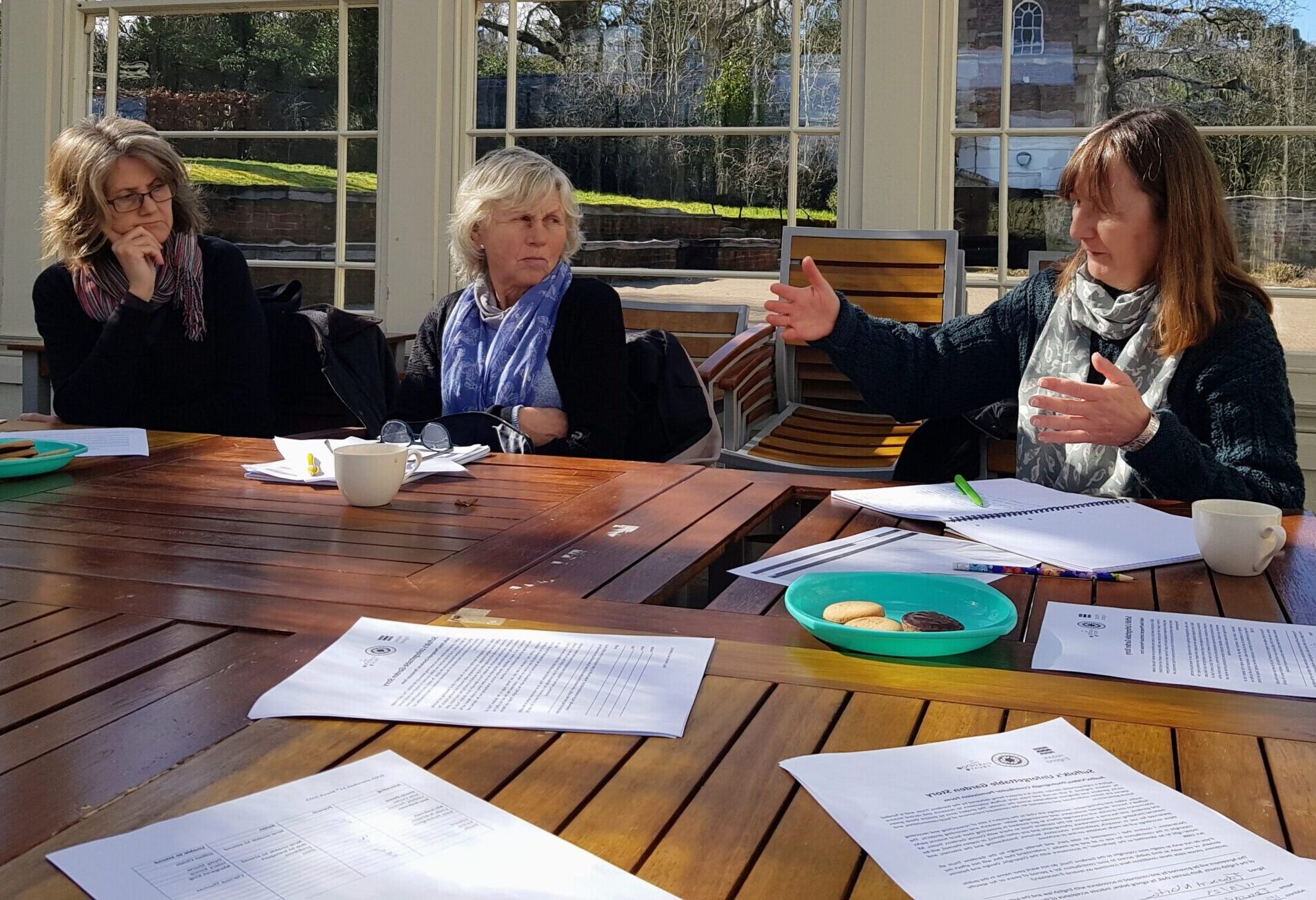 Suffolk based research volunteers sitting around a table with papers of their research, engaged in discussion inside a sunny outdoor glasshouse