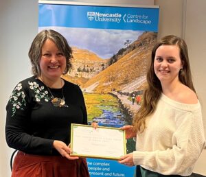 Two women wearing jumpers, one white and the other a black jumper, smiling and jointly holding a white certificate in a gold frame.