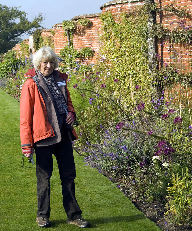 Susan Campbell standing in an orange jacket by a red brick crinkle-crankle wall lined with a flower bed