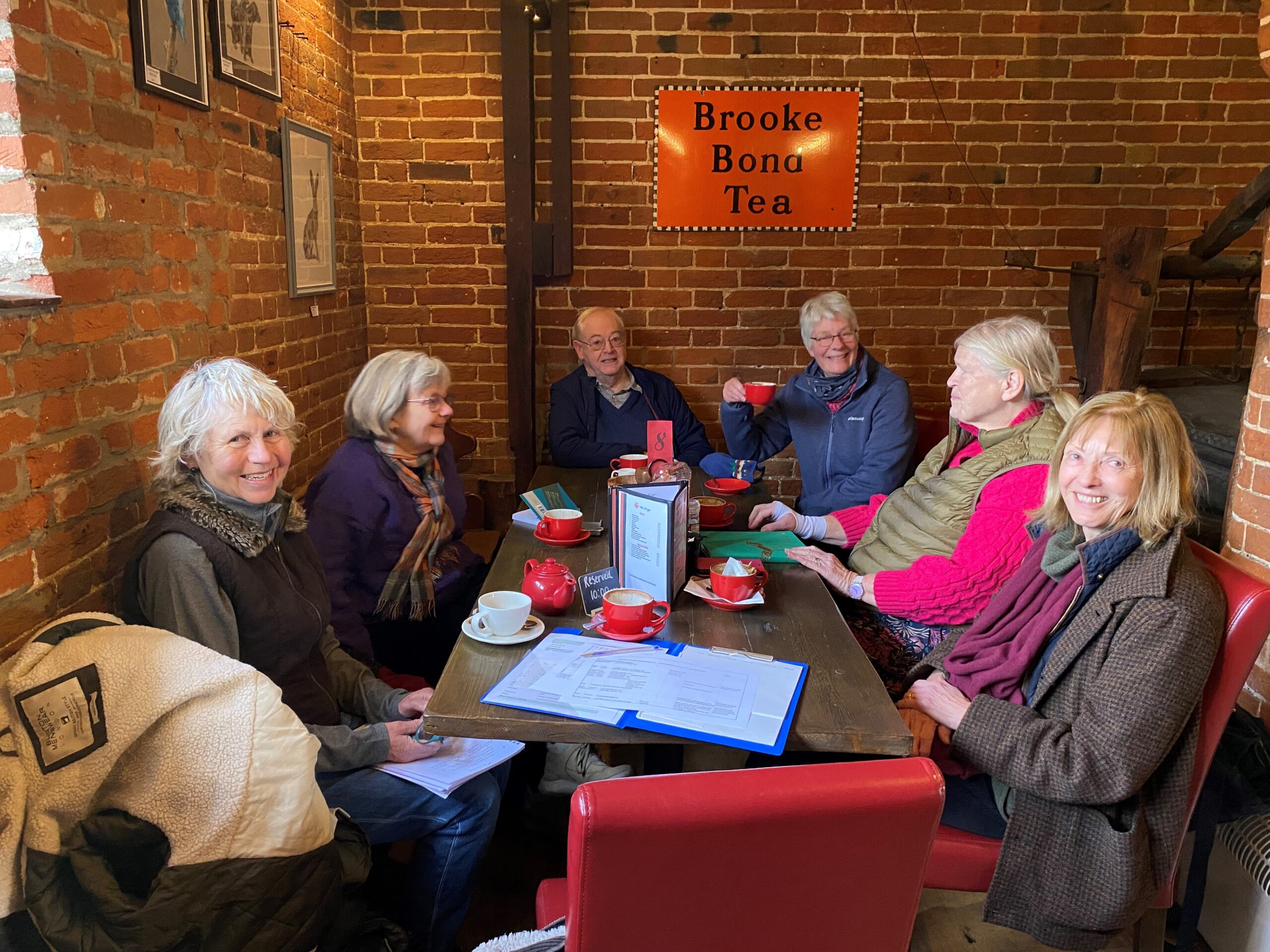 Group of 6 volunteers sat around a table, drinking tea in a cosy cafe in Suffolk with red brick wall background.