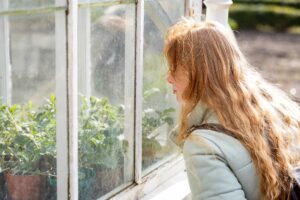 Woman with long ginger hair looking through a glasshouse at green leafy plants.