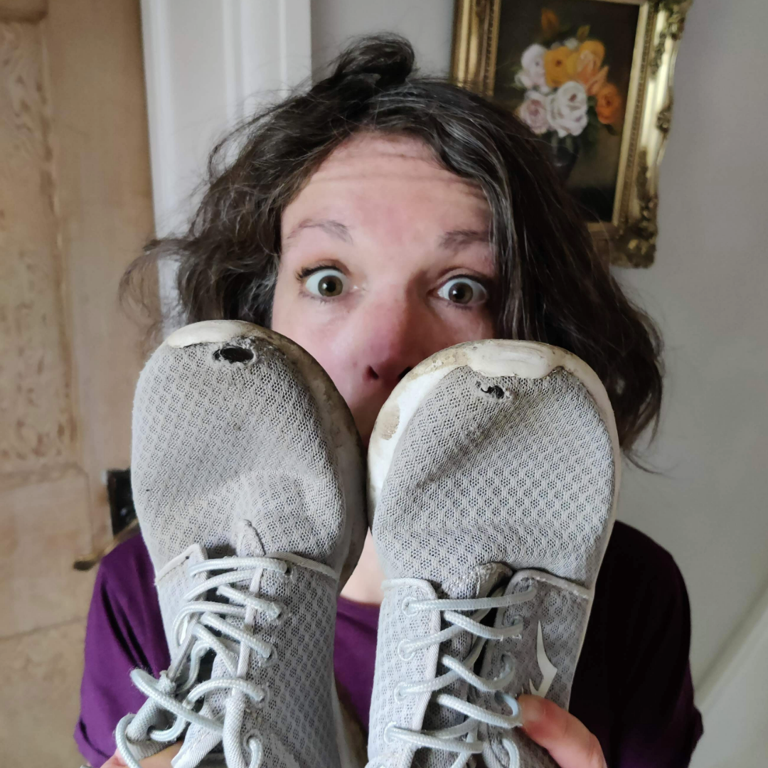 Linden holds up a pair of worn grey trainers with holes in the toes, covering half her surprised face.