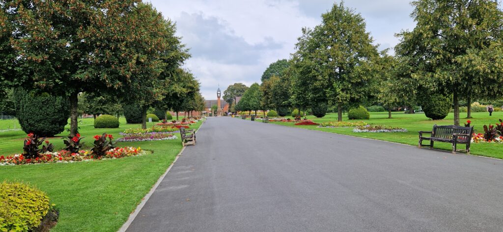 View of main path in Queens Park, Crewe, in summer, bordered with leafy green trees, benches and colourful bedding plants.