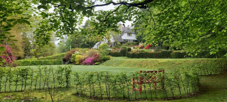 Lush green gardens of arts and crafts house High Moss, with pink and red plants and green leafy trees and a red bench nestled amongst bushes in the foreground.