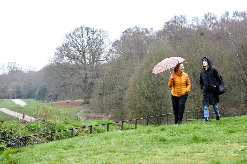 Two women, one holding an umbrella in a yellow coat the other in a black hooded coat, walking through a park in the rain.