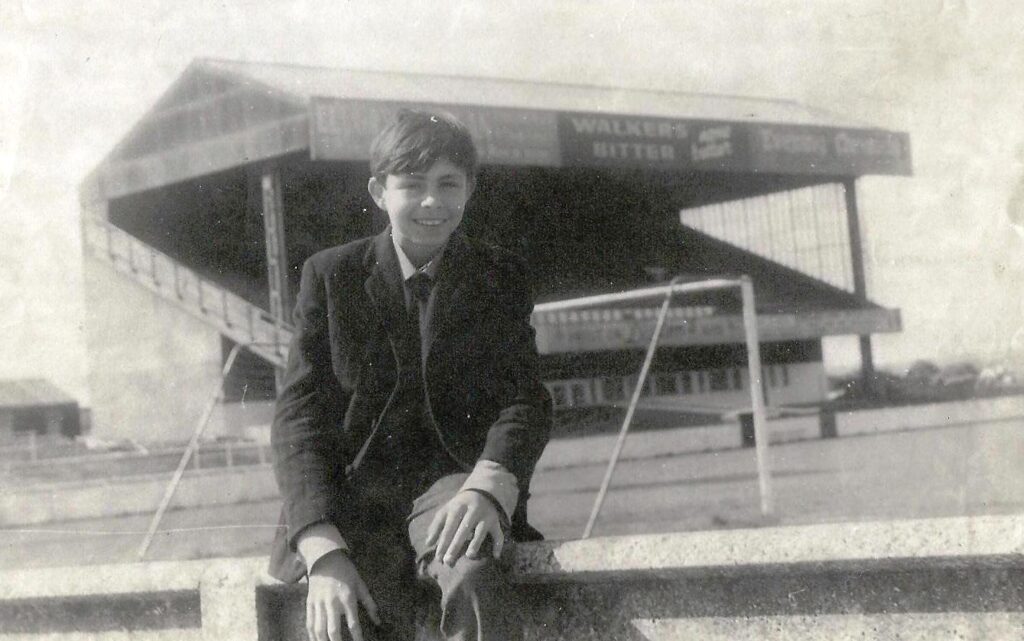 Black and white photograph of a young boy, John Harris, sat in front of a football stand in 1968 at Springfield Park, Wigan Athletic