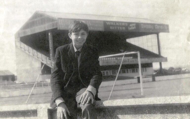 Black and white photograph of a young boy, John Harris, sat in front of a football stand in 1968 at Springfield Park, Wigan Athletic